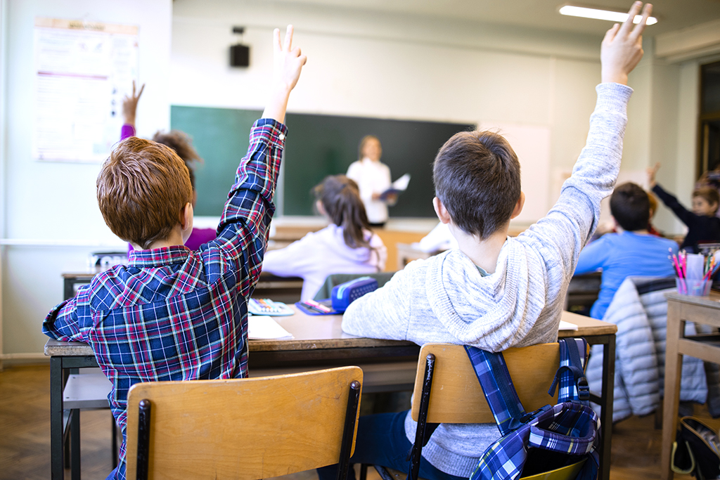 Schoolchildren at classroom with raised hands answering teacher's question.